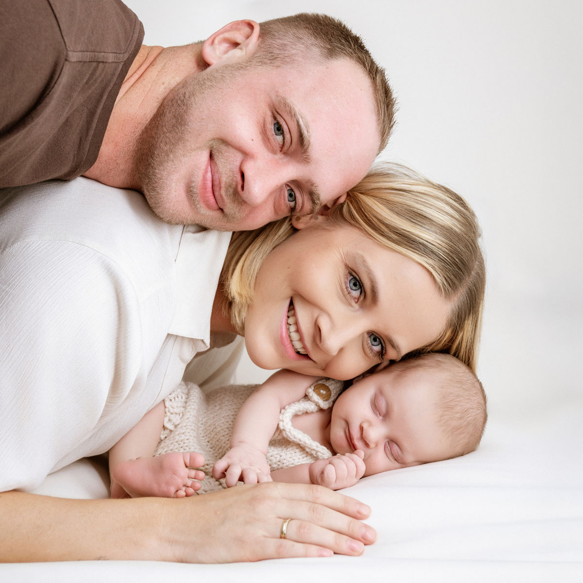 Mama und Papa liegen lächelnd neben ihrem schlafenden Neugeborenen