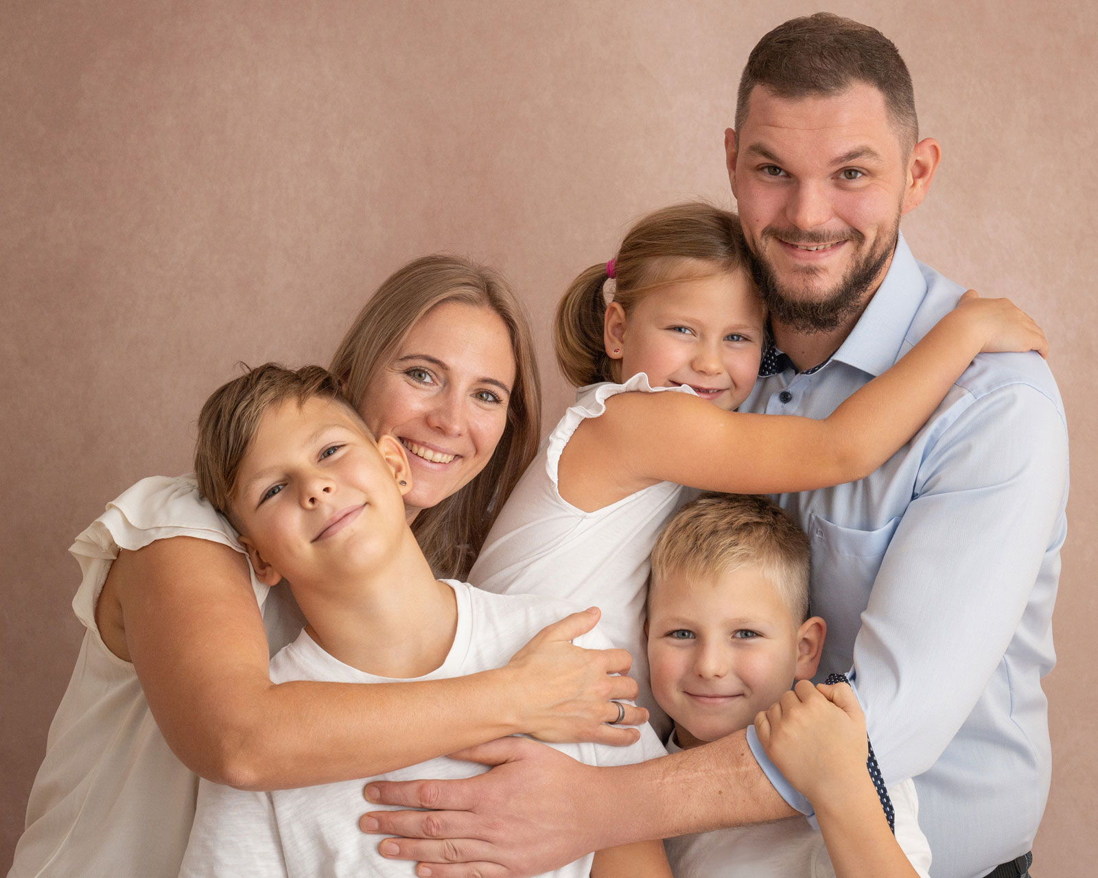 Familie mit drei Kindern umarmt sich lachend vor einem beigefarbenen Hintergrund